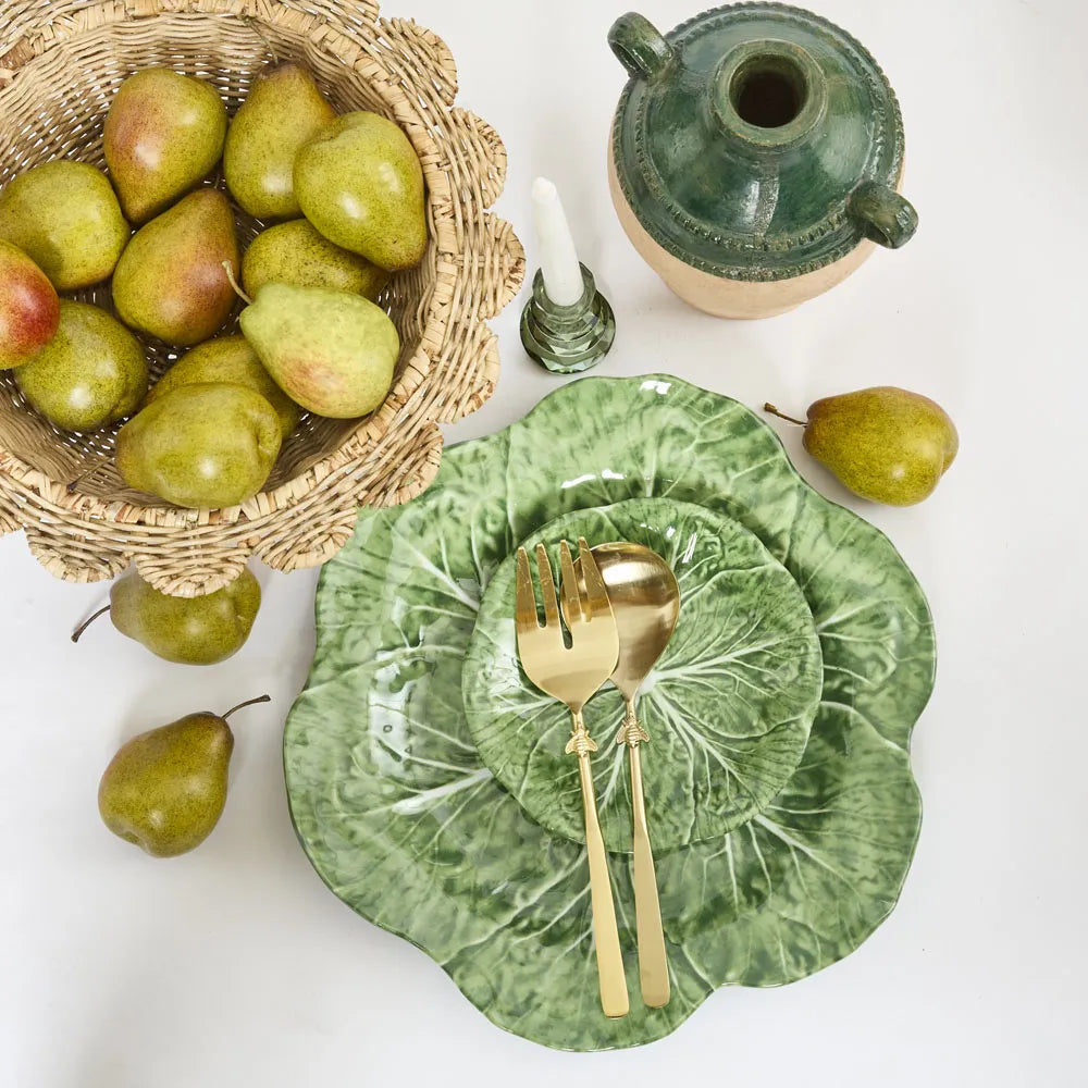 Green ceramic plates with gold cutlery, a basket of pears, and a green ceramic container on a white surface.