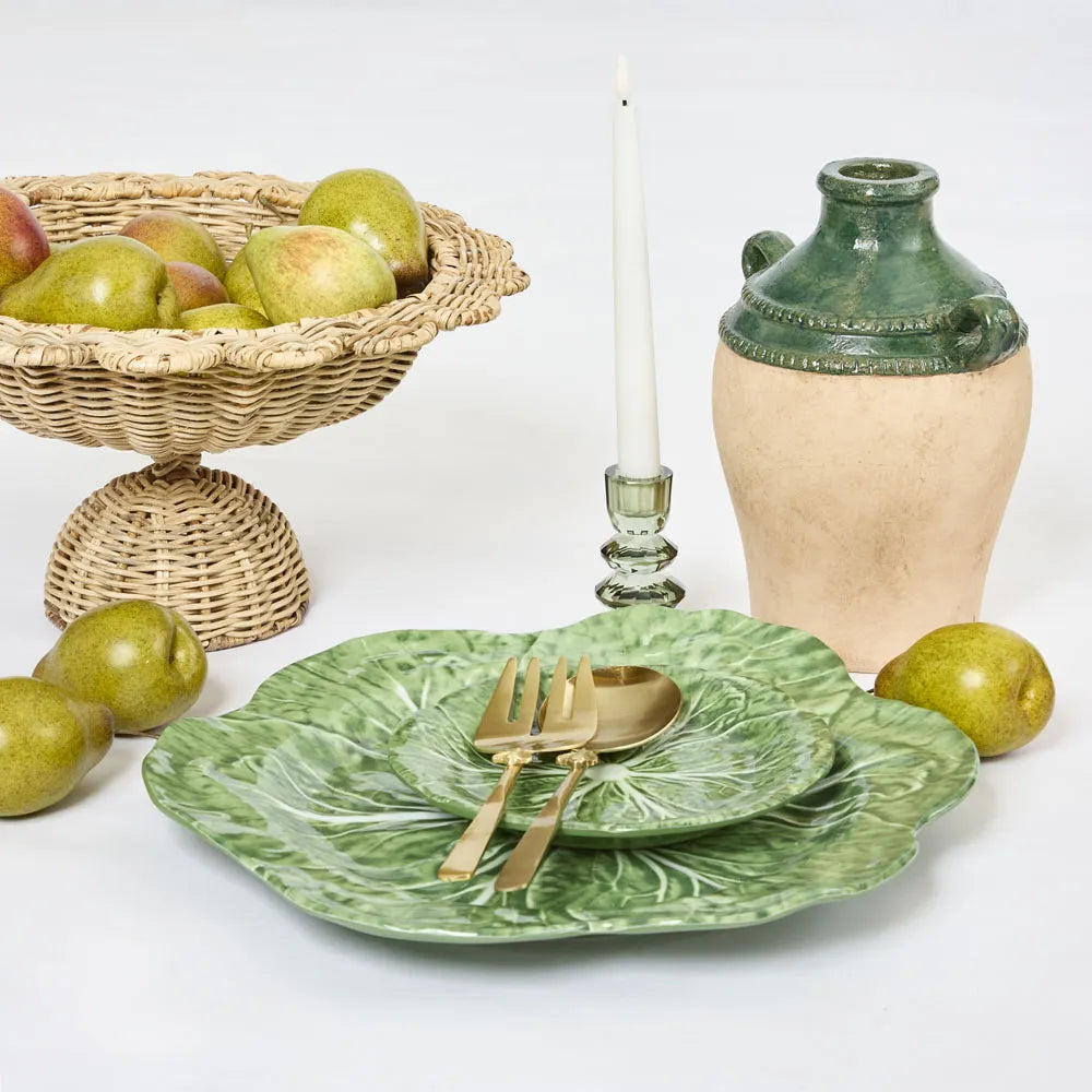 Green ceramic plates with gold cutlery, a woven basket of apples, and a ceramic vase on a white background.