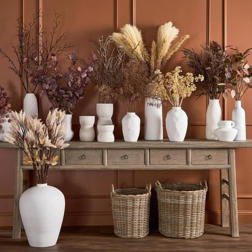 Decorative setup with vases, bowls, and dried plants on a wooden console table against a brown paneled wall.